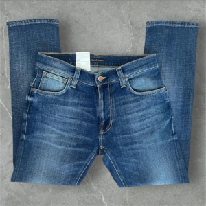 Blå jeans från Nudie Jeans - Snygga blå jeans från Nudie Jeans med klassisk femficksdesign och subtila slitningar. De har en normal passform och är perfekta för en avslappnad stil. Jeansen har en knappgylf och är tillverkade i högkvalitativt denim. Helt nya aldrig använda.