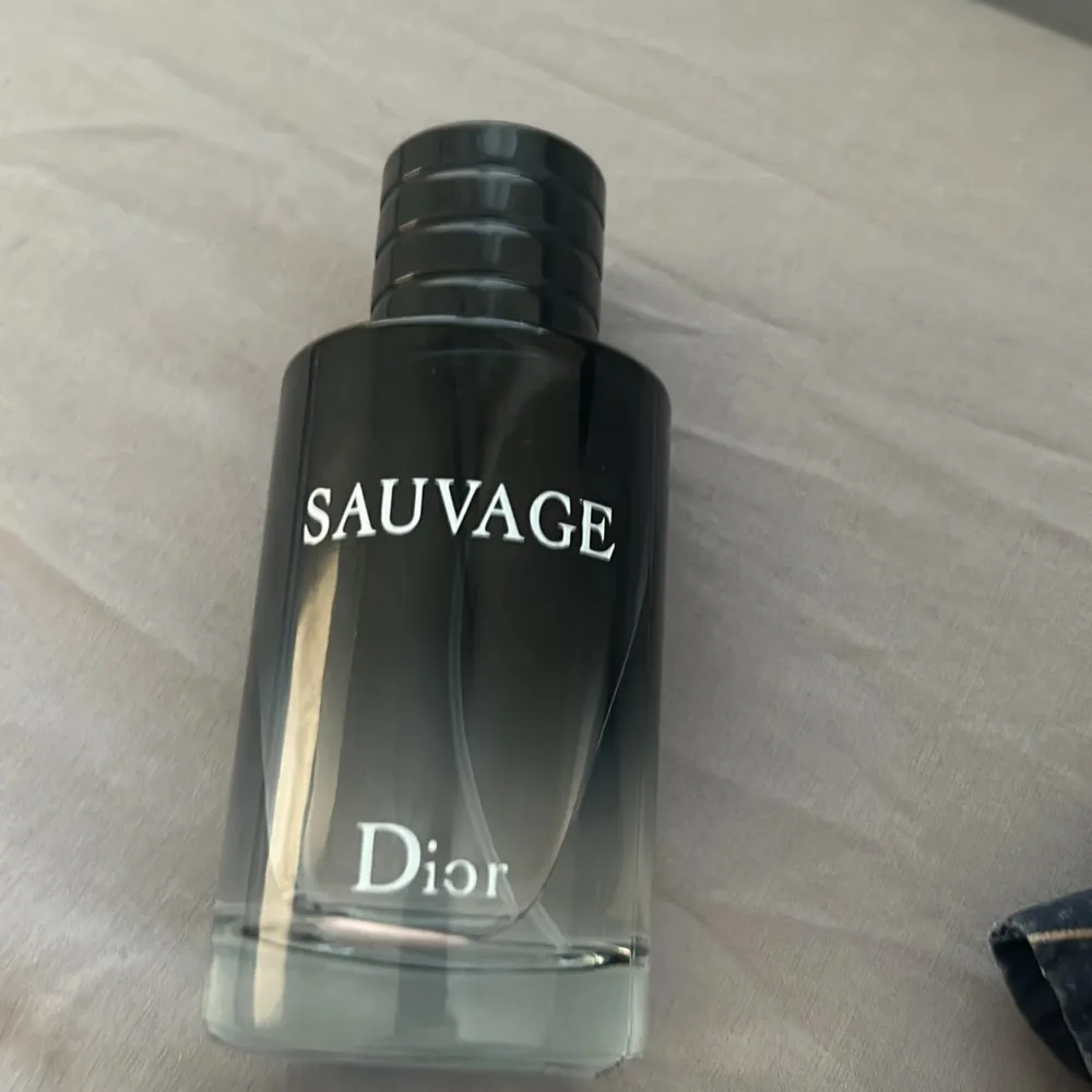 Säljer en elegant flaska av Sauvage parfym från Dior. Flaskan har en stilren design med en mörk toning som övergår till klar vid botten. Korken är svart med Diors logotyp på toppen. Perfekt för den som vill ha en tidlös och maskulin doft.. Perfume.