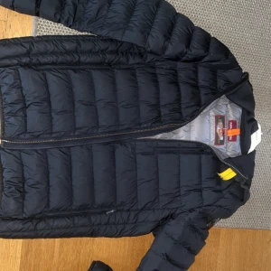 Svart dunjacka från Parajumpers med luva  - Säljer en svart dunjacka från Parajumpers med en lätt och bekväm design. Jackan har en dragkedja framtill och en broderad logga på ärmen. Perfekt för kyliga dagar med stil.