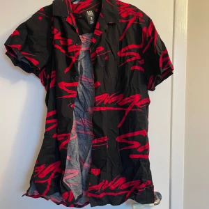 Svart och röd kortärmad skjorta från Black Squad - Snygg kortärmad skjorta från Black Squad i svart med röda abstrakta mönster. Perfekt för en avslappnad stil med knappar framtill och en klassisk krage.
