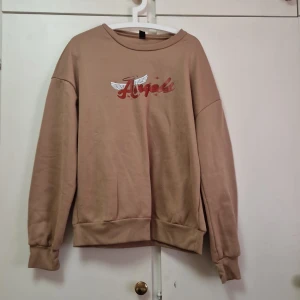 Beige sweatshirt med tryck - Säljer en beige sweatshirt med texten 'Angel' i rött och vita vingar på bröstet. Tröjan har långa ärmar och ribbade muddar vid ärmslut och nederkant. Perfekt för en avslappnad stil.