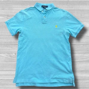 Ralph Lauren piké - Snygg blå pikétröja från Polo Ralph Lauren i slim fit. Tröjan har en klassisk krage med knappar och det ikoniska broderade logotypen i gult på bröstet. Perfekt för en stilren look.