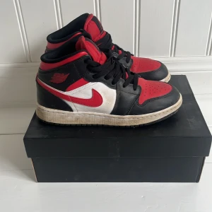 Nike Air Jordan 1 Mid (GS) i svart, röd och vit - Säljer ett par Nike Air Jordan 1 Mid (GS) i klassiska färgerna svart, röd och vit. Skorna har en ikonisk design med snörning och rund tå. Perfekta för sneakerheads som älskar en tidlös stil.