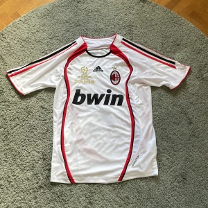 Vit AC Milan fotbollströja från Adidas - Säljer en vit retro AC Milan fotbollströja från Adidas med röda och svarta detaljer. Tröjan har korta ärmar och klubbmärket på bröstet. Namnet 'Maldini' och nummer 3 är på baksidan. Tröjan är från finalen 2007 i Aten. Nypris: 550 kr! 