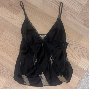 Svart linne med spetsdetaljer från na-kd - Elegant svart linne från NA-KD med smala axelband och spetsdetaljer. Toppen har ett subtilt mönster och en v-ringad design som ger en stilren look. Perfekt för en kväll ute eller en speciell tillställning.