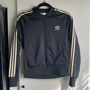 Svart och guldig tröja från Adidas - Snygg svart tröja från Adidas med guldiga ränder längs ärmarna och Adidas-logga på bröstet. Tröjan har dragkedja framtill och långa ärmar. Meshfoder så lite tjockare. 