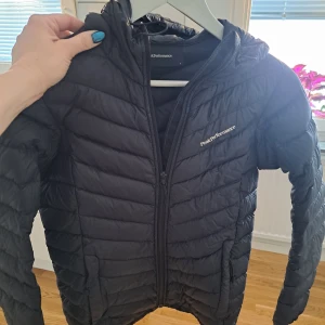 Svart dunjacka från Peak Performance - Säljer en svart dunjacka från Peak Performance. Jackan har en dragkedja framtill och är quiltad för extra värme. Den har en huva och långa ärmar, perfekt för kyligare dagar. Finns några små hål bak och den är ihopsydd i en söm .syns om man tittar nära annars inte på håll 