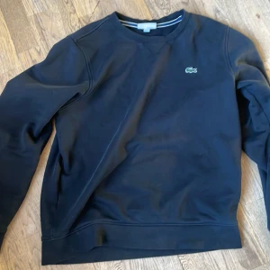 Svart sweatshirt från Lacoste Sport - Säljer en stilren svart sweatshirt från Lacoste Sport med det klassiska krokodilmärket på bröstet. Tröjan har några mindre slitage på den högra ärmen.
