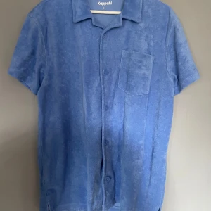Blå frottéskjorta från Kappahl - Säljer en blå kortärmad frottéskjorta från Kappahl. Skjortan har en klassisk krage, knappar framtill och en bröstficka. Perfekt för en avslappnad stil.
