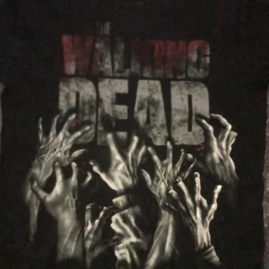 Svart långärmad tröja med The Walking Dead-tryck - Säljer en svart långärmad tröja med The Walking Dead-tryck. Tröjan har ett coolt motiv av händer och texten 'INFECTED' på ärmen. Perfekt för fans av serien!