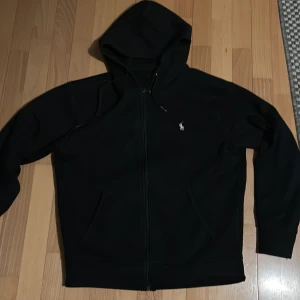 Svart hoodie från Polo Ralph Lauren - Säljer en stilren svart hoodie från Polo Ralph Lauren. Den har en dragkedja framtill och en liten logotyp på bröstet. Perfekt för en avslappnad look. 🖤