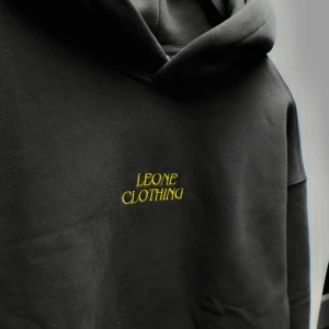 Svart hoodie från Leone Clothing (M,L,XL) - Snygg svart hoodie från Leone Clothing med guld text på framsidan och ett lejonmotiv på baksidan. Perfekt för en casual look med sin bekväma passform och klassiska design. Kan användas i vardagen eller till och från gymmet. Stötta LeoneClothing genom att slå till idag! Finns i flera olika storlekar.