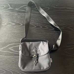 Svart axelväska från Trapstar - Snygg svart axelväska från Trapstar med justerbar axelrem och coolt spänne framtill. Väskan har en dragkedja upptill och är perfekt för att bära många saker då den har mycket utrymme.