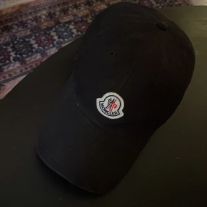 Svart keps från Moncler - Snygg svart keps från Moncler med deras ikoniska logotyp framtill. Kepsen har en justerbar rem baktill med röd och vit detalj för perfekt passform. Perfekt för en stilren look.