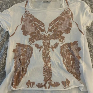 Vit t-shirt med brunt mönster - Säljer en vit t-shirt med ett unikt brunt mönster framtill. T-shirten har korta ärmar och är tillverkad i ett lätt och bekvämt material. 