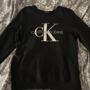 Svart tröja från Calvin Klein Jeans - Snygg svart tröja från Calvin Klein Jeans med stort vitt logotryck på framsidan. Perfekt för en stilren och avslappnad look. Tröjan har långa ärmar och rund halsringning.