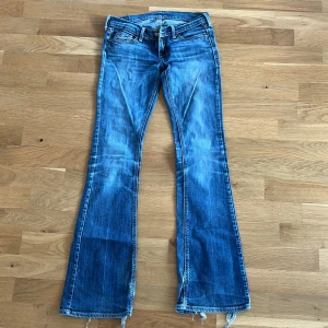 Blå bootcut jeans - Snygga blå bootcut jeans med slitna detaljer och fransar vid bensluten. Jeansen har en klassisk femficksdesign och en låg midja. Perfekta för en avslappnad stil.