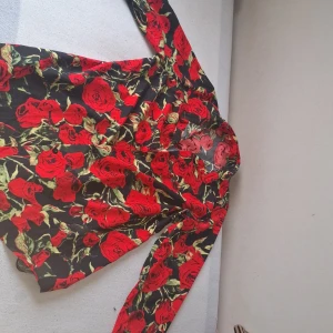 Svart skjorta med röda rosor - Säljer en svart skjorta med stora röda rosor och gröna blad. Skjortan har lång ärm, klassisk krage och knäppning framtill med svarta knappar. Perfekt för dig som vill sticka ut med ett färgstarkt mönster. Perfekt för en sommar härlig kännsla