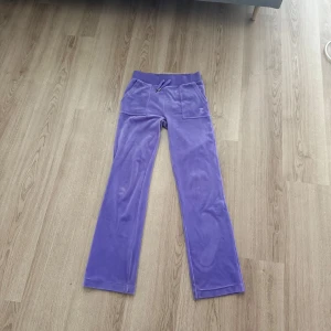 Lila velourbyxor från Juicy Couture - Säljer ett par lila velourbyxor från Juicy Couture i storlek XS. Byxorna har raka ben, resår och snörning i midjan samt fickor både fram och bak med knappdetaljer. Perfekta för en avslappnad och trendig look.