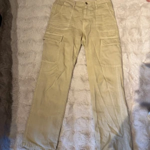 Beige cargobyxor med fickor - Säljer ett par beige cargobyxor med flera stora fickor på sidorna och bak. Byxorna har rak passform. En knapp på ena sidfickan har lossnat men det är inget som syns eller påverkar användningen