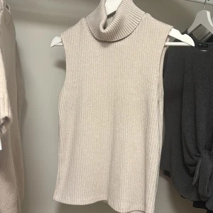 Beige ribbad ärmlös polotopp från Zara - Säljer en beige ribbad polotopp från Zara. Toppen är ärmlös med hög polokrage och har en mjuk, stickad känsla. Perfekt att styla lager på lager eller bära som den är.