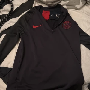 Svart Paris Saint-Germain träningströja från Nike - Säljer en svart långärmad träningströja från Nike med Paris Saint-Germain-logga på bröstet. Tröjan har halv dragkedja och röda detaljer, inklusive Nike-loggan och inslag vid kragen. Perfekt för fotbollsträning.