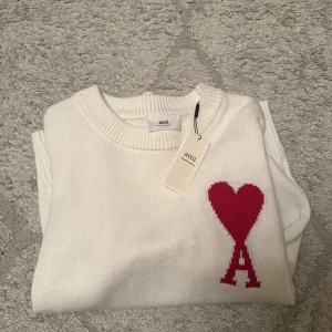 Vit stickad tröja med rött hjärta från Ami - Vit stickad tröja från Ami med rund halsringning och ett rött hjärta med bokstaven A broderat på bröstet. Tröjan har långa ärmar och ribbade muddar vid ärmslut och nederkant. Perfekt för en stilren och trendig look. Helt ny. Kontakta för mer info o bilder