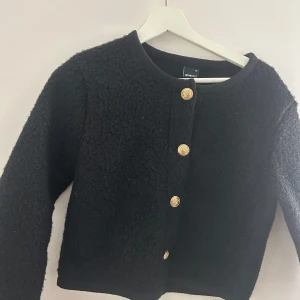 Svart cardigan jacka med guldknappar från Gina Tricot - Säljer en svart cardigan jacka från Gina Tricot med rund halsringning och fyra dekorativa guldknappar framtill. Jackan har lång ärm och är i ett mjukt, strukturerat material. Perfekt att slänga över en topp för en snygg look.