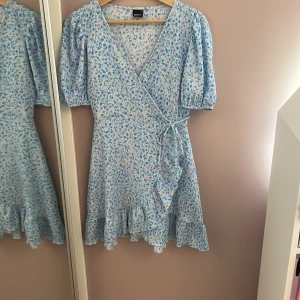 Blommig omlottklänning från Gina Tricot - Söt ljusblå och vit omlottklänning från Gina Tricot med blommigt mönster. Klänningen har puffiga korta ärmar, volangkant nertill och knyt i midjan. Perfekt för vår och sommar! Använd endast 1 gång och är därför i ny skick! 