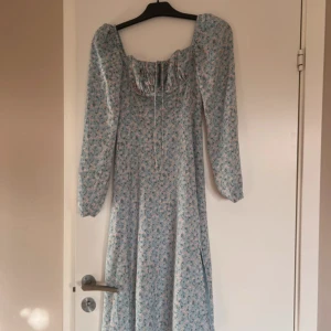 Blommig långklänning från Adoore - Supersöt långklänning från Adoore i ljusblått med små blommor. Klänningen har långa puffärmar, fyrkantig ringning och dragkedja i ryggen. Perfekt för vår och sommar!