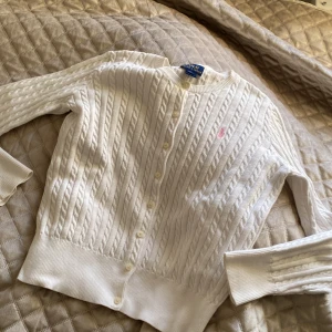 Vit kabelstickad kofta från Polo Ralph Lauren - Säljer en klassisk vit kabelstickad kofta från Polo Ralph Lauren med rund halsringning och knappar framtill. Tröjan har lång ärm och en liten rosa broderad logga på bröstet. 