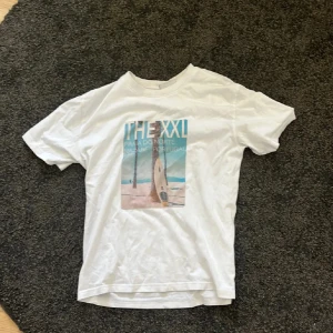 Vit grunt T-shirt i storlek L i barnstorlek  - Vit t-shirt från Grunt i storlek L med tryck framtill som visar en surfbräda på stranden och texten 'THE XXL'. T-shirten har rund hals och korta ärmar. Perfekt för en avslappnad stil.