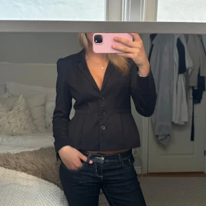 Svart kavaj med markerad midja - Säljer en svart kavaj med figurnära passform och markerad midja. Kavajen har två knappar framtill, klassisk krage och långa ärmar. Perfekt för en stilren och dressad look.