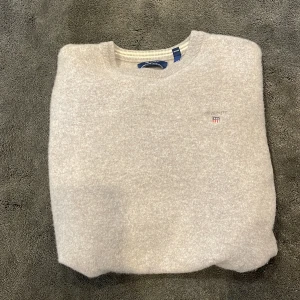 Ljusgrå ulltröja från GANT - Säljer en stilren ljusgrå tröja från GANT i superfin lammull🐑. Tröjan har rund halsringning, ribbade muddar och en liten broderad GANT-logga på bröstet. Perfekt för dig som gillar klassisk och enkel stil. Skriv gärna vid frågor