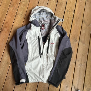 Grå och svart vindjacka från Mammut - Säljer en grå och svart vindjacka från Mammut med huva och dragkedja. Jackan har två bröstfickor med dragkedja och justerbar nederkant. Perfekt för utomhusaktiviteter och har en sportig look.