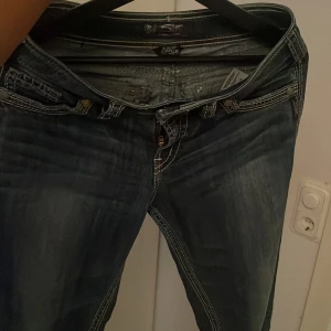 Silver Jeans Suki bootcut jeans - Säljer ett par blå bootcut jeans från Silver Jeans, modell Suki. Jeansen har snygga kontrastsömmar och broderade bakfickor. Klassisk femficksmodell med låg midja och dragkedja. Perfekta för dig som gillar western-inspirerad stil.