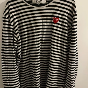 Randig långärmad tröja från Comme des Garçons Play - Säljer en svartvit randig långärmad tröja från Comme des Garçons Play i storlek XL men passar L okså jättebra. Tröjan har ett rött hjärta med ögon broderat på bröstet och är tillverkad i bomull. Perfekt för dig som gillar stilrena och ikoniska plagg.
