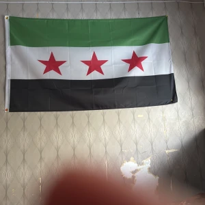 Syriska oppositionsflaggan - Säljer en tygflagga med tre röda stjärnor på vit bakgrund, med grönt fält upptill och svart fält nedtill. Flaggan är rektangulär och har metallöljetter i hörnen för upphängning.