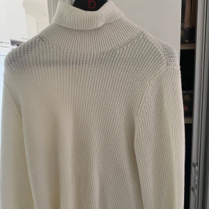 Vit stickad polotröja - Säljer en stilren vit stickad polotröja med lång ärm. Tröjan har en hög krage och är ribbstickad i både krage och muddar. Perfekt för lager-på-lager och passar till många olika outfits.