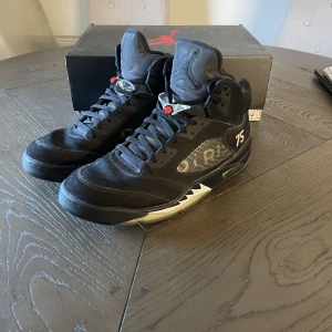 Jordan 5 PSG - Säljer ett par svarta Nike Air Jordans PSG med grå och vita detaljer. Skorna har nummer 75 på sidan, PSG-logga på hälen och genomskinlig sula med röd PSG-grafik undertill. Klassisk hög modell med snörning och rund tå.