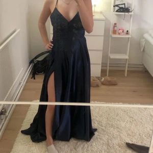 Mörkblå långklänning med slits från JJ's House - Säljer en elegant mörkblå långklänning från JJ's House i glansigt satinmaterial. Klänningen har djup v-ringning, smala axelband med korsad snörning i ryggen och hög slits framtill. Perfekt för bal eller festliga tillfällen. Den är i nyskick och har aldrig använts bara testat klänningen en gång, super fin men kände inte att den skulle komma till användning!