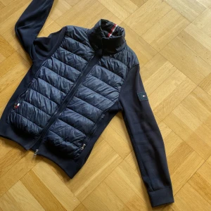 Mörkblå cardigan, Tommy Hilfiger - Snygg mörkblå hybridjacka från Tommy Hilfiger med quiltad front och rygg i glansigt material samt stickade ärmar. Jackan är som ny, köptes för 2700kr. Perfekt för lager-på-lager och stilren look 🥇