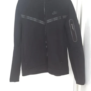 Nike Tech Hoodie - Svart träningsjacka från Nike med dragkedja och diskret logga på bröstet. Jackan har lång ärm och en ficka med dragkedja på ena ärmen. Snygga detaljer med svarta band över bröstet.
