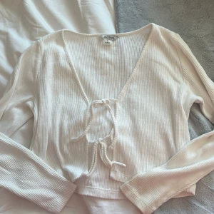 Vit ribbad knyt-topp från Monki - Säljer en vit ribbad topp från Monki med lång ärm och v-ringning. Toppen har snygga knytband framtill och är croppad i modellen. Perfekt för att styla med jeans eller kjol.
