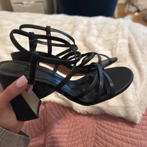 Svarta klackade sandaler från H&M - Säljer ett par svarta klackar från H&M. Skorna har tunna remmar som korsar över foten och runt vristen, samt en öppen tå. Perfekta för sommarens tillfällen. Har använts ett fåtal gånger