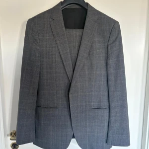 Grå rutig kostymsett från BOSS - Säljer ett stilrent grå rutig kostymsett från BOSS. Kavajen har klassisk passform med två knappar och byxorna har raka ben. Perfekt för dig som vill ha en tidlös och elegant look.