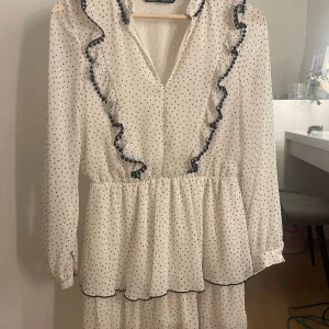 Vit prickig klänning med volang från Zara - Supersöt vit klänning från Zara med svarta prickar och volangdetaljer längs v-ringningen. Långärmad modell med markerad midja och lätt volang i kjolen. Perfekt för skolavslutning, student, konfirmation osv. Säljer eftersom dem är för liten🩷