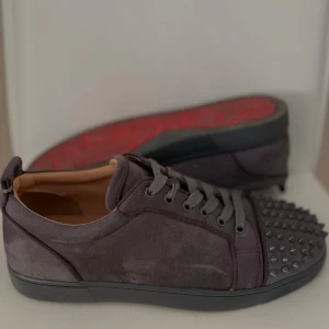 Gråa sneakers med nitar från Christian Louboutin - Säljer ett par gråa sneakers från Christian Louboutin med ikonisk röd sula och coola nitar framtill. Skorna har snörning och är tillverkade i mocka med detaljer i skinn. Perfekta för dig som vill sticka ut med din stil. Smutsiga på bilderna men putsas före leverans. Box och dust ingår. Ifall fler frågor vill ställas så kan man komma via dm!