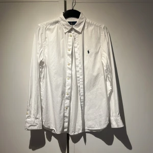 Vit skjorta från Ralph Lauren - Klassisk vit skjorta från Ralph Lauren med broderad logga på bröstet. Skjortan har lång ärm, knappar framtill och krage. Perfekt för en stilren och tidlös look.