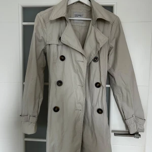 Beige trenchcoat från Esprit - Snygg beige trenchcoat från Esprit med klassisk dubbelknäppning och bälte i midjan. Jackan har långa ärmar, bred krage och knappar vid ärmslut. Perfekt för övergångssäsonger och ger en stilren look. Har en liten fläck på ärmen som syns på sista bilden. Ungefär 77 cm lång 